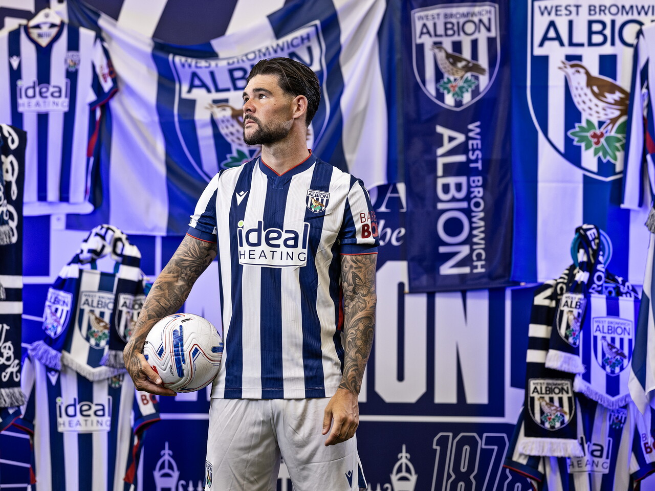 Alex Mowatt in the home kit holding a ball while stood in front of an Albion back drop with plenty of WBA branding