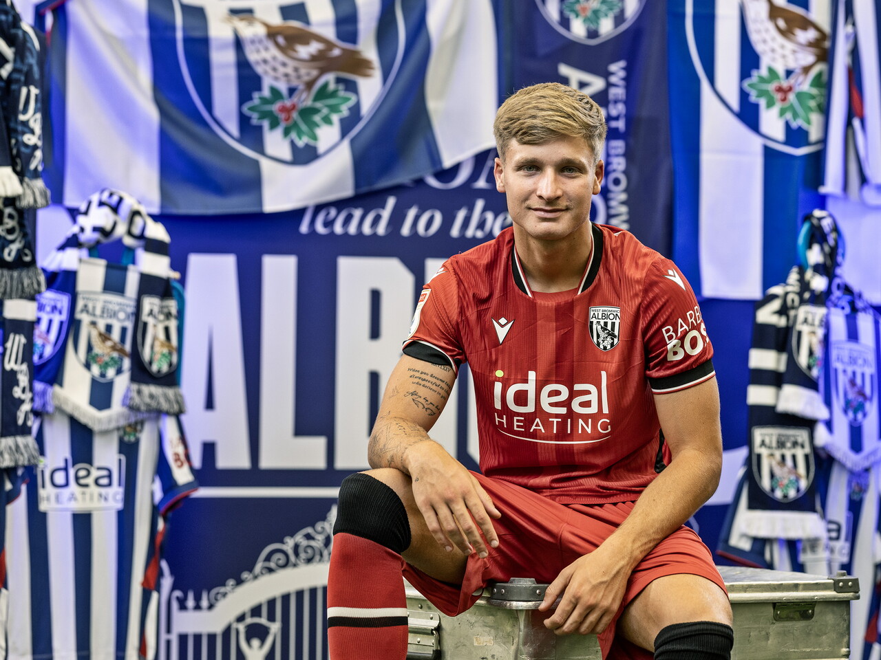 JJ Morgan in the red away kit smiling at the camera while sat in front of an Albion back drop with plenty of WBA branding