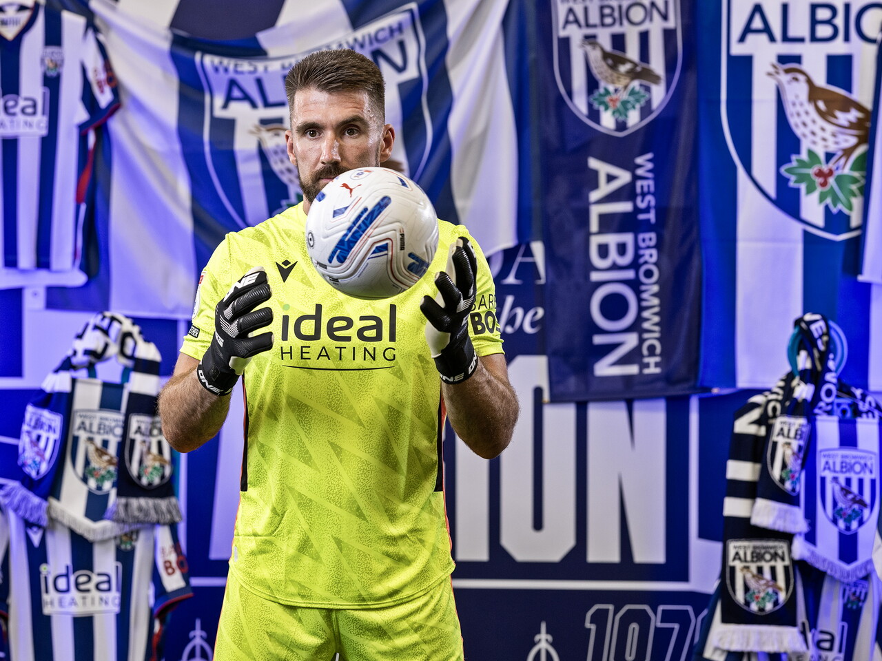 Matt Ingram in the yellow GK kit looking at the camera and holding a ball while stood in front of an Albion back drop with plenty of WBA branding