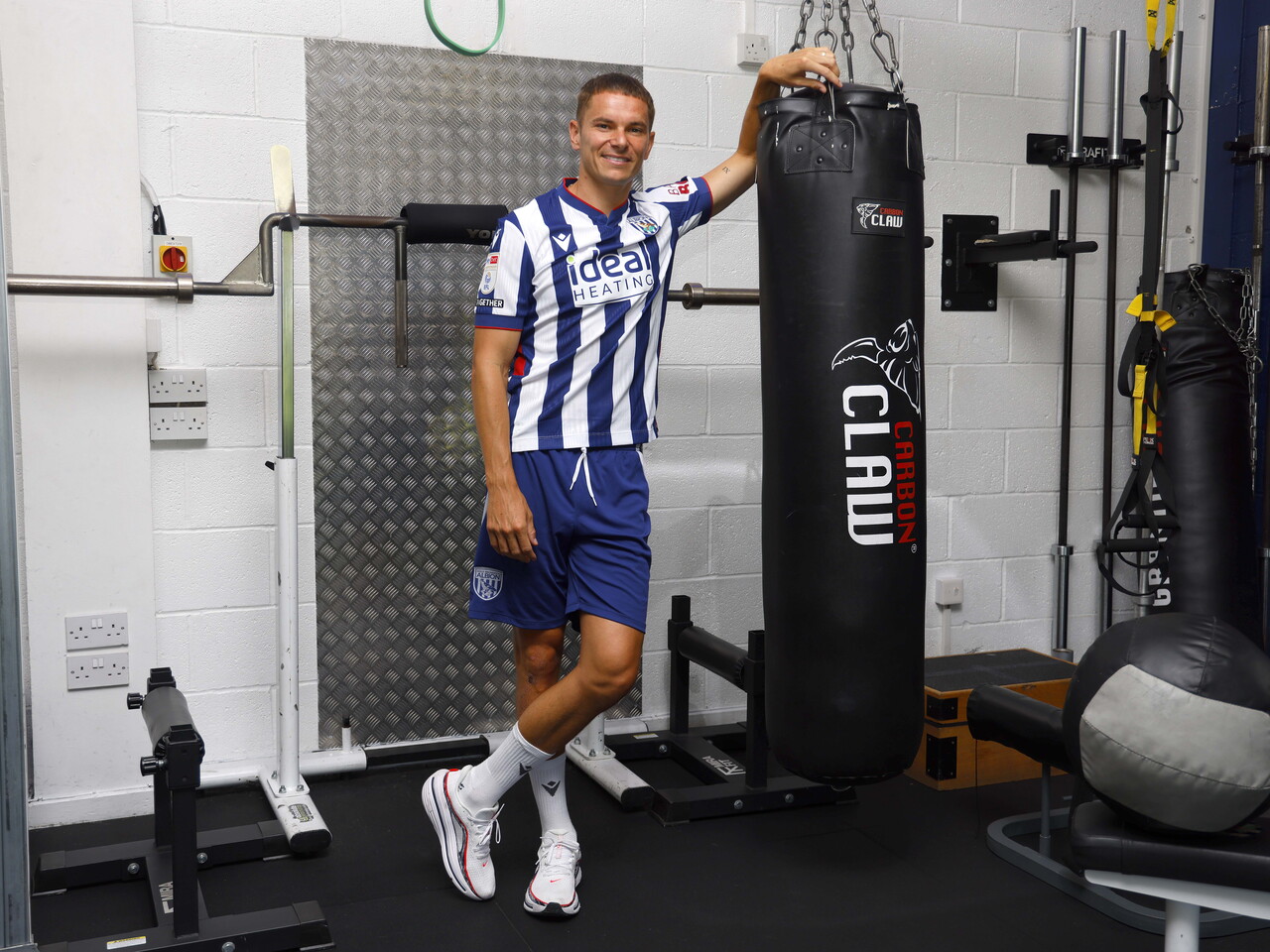 Conor Townsend smiling at the camera while leaning up against a punch bag in the gym while stood in a home shirt 