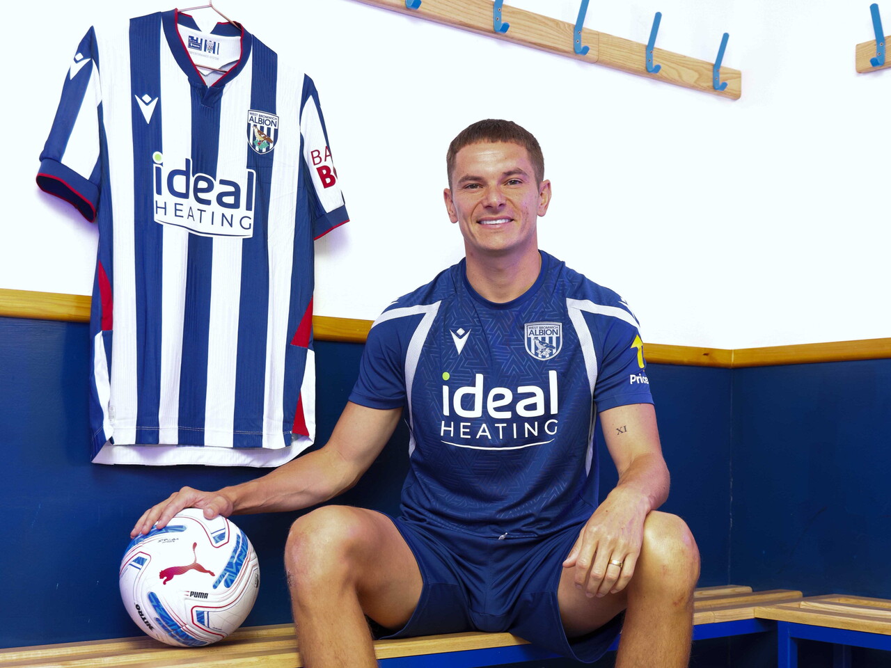 Conor Townsend smiling at the camera while sat on a bench in the dressing room with his hand on a football and a home shirt hanging up behind him
