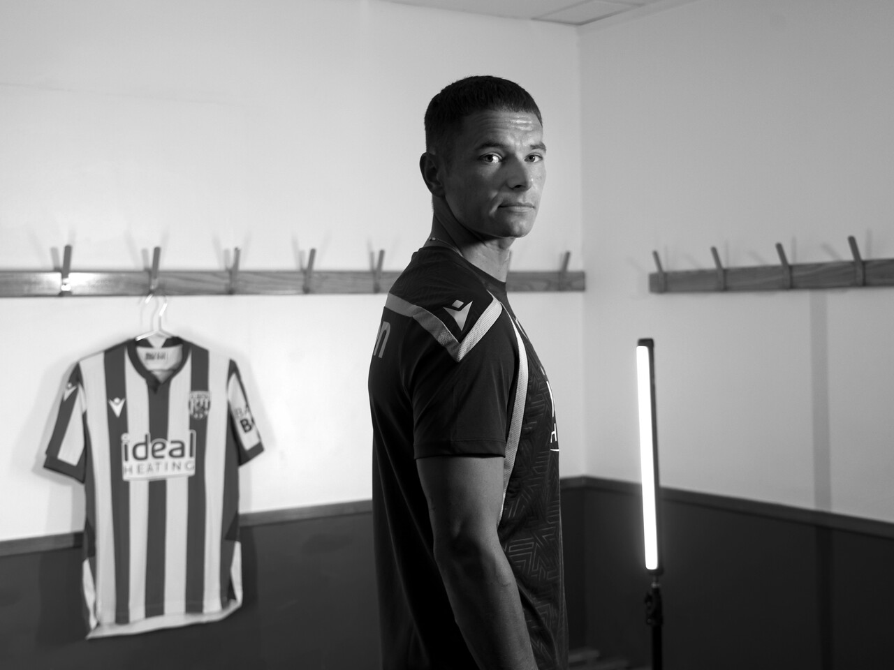 Conor Townsend looking at the camera stood side-on in a dressing room with a home shirt hanging up behind him