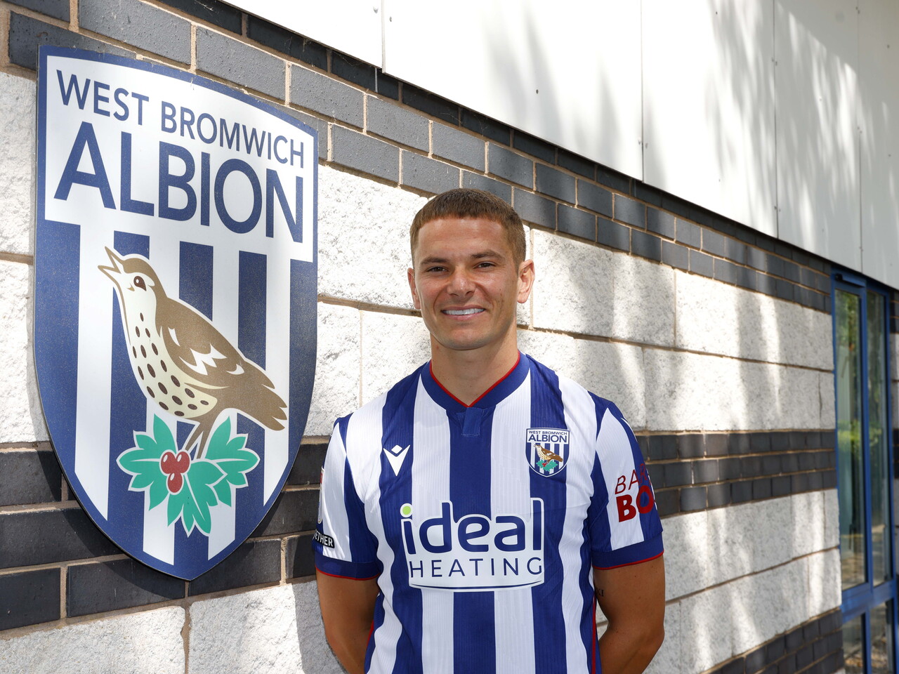 Conor Townsend smiling at the camera while stood in the home shirt next to a big WBA badge on a wall