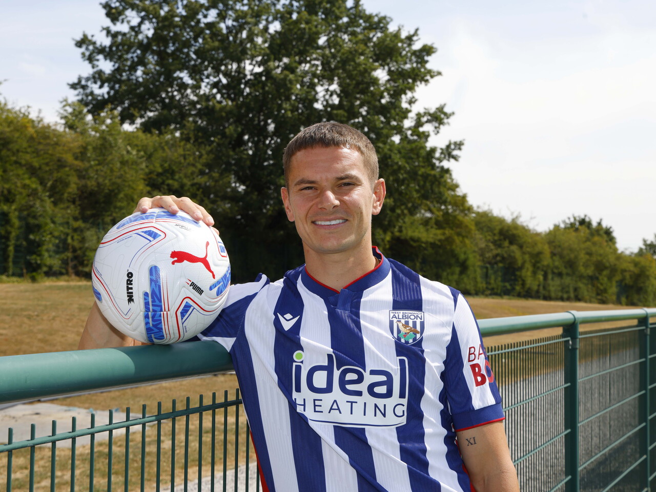 Conor Townsend smiling at the camera while stood in the home shirt and holding a ball, leaning up against a fence