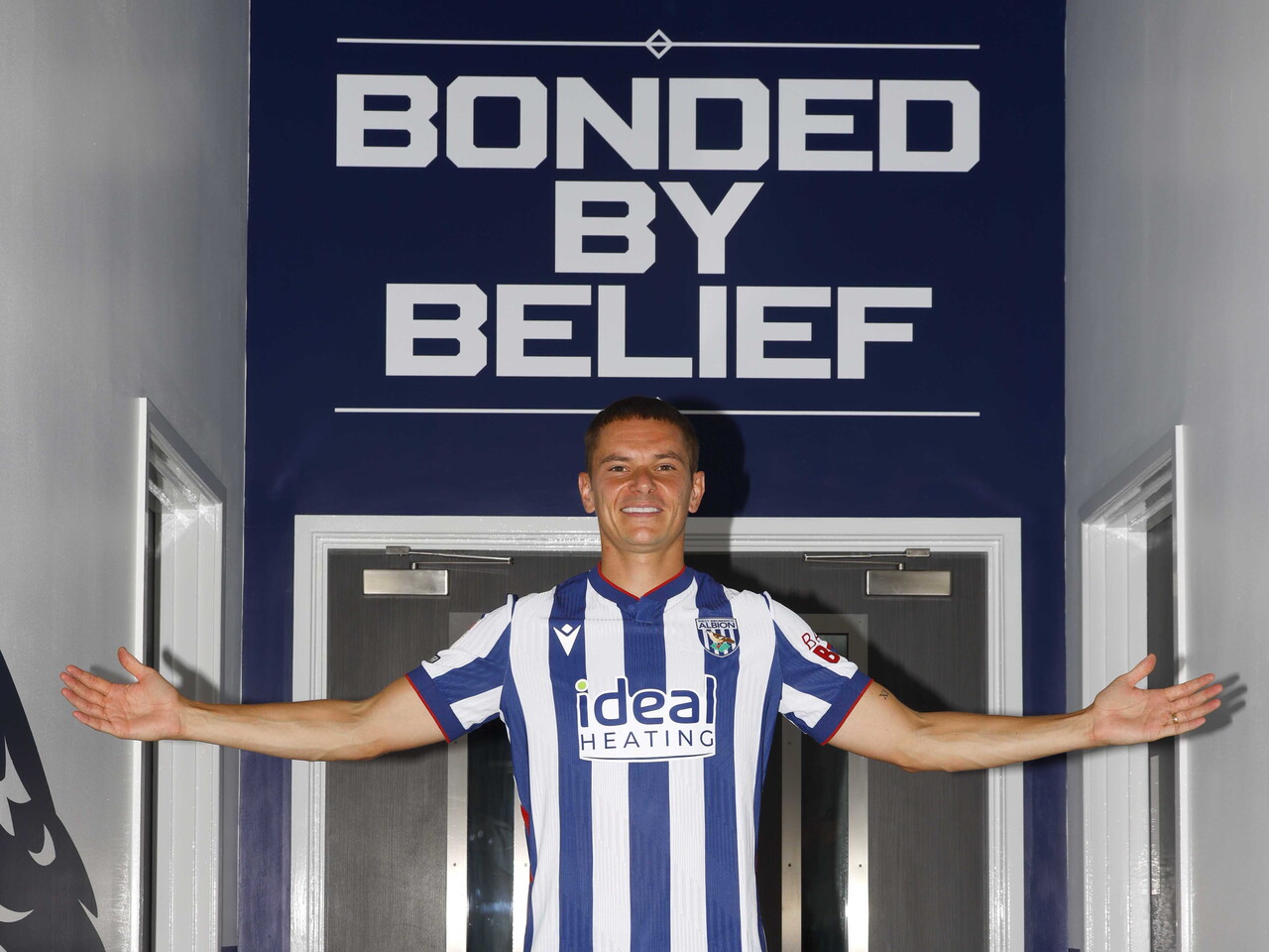 Conor Townsend with his arms outstretched smiling at the camera while stood in the home shirt in front of branding which reads 'Bonded by Belief'
