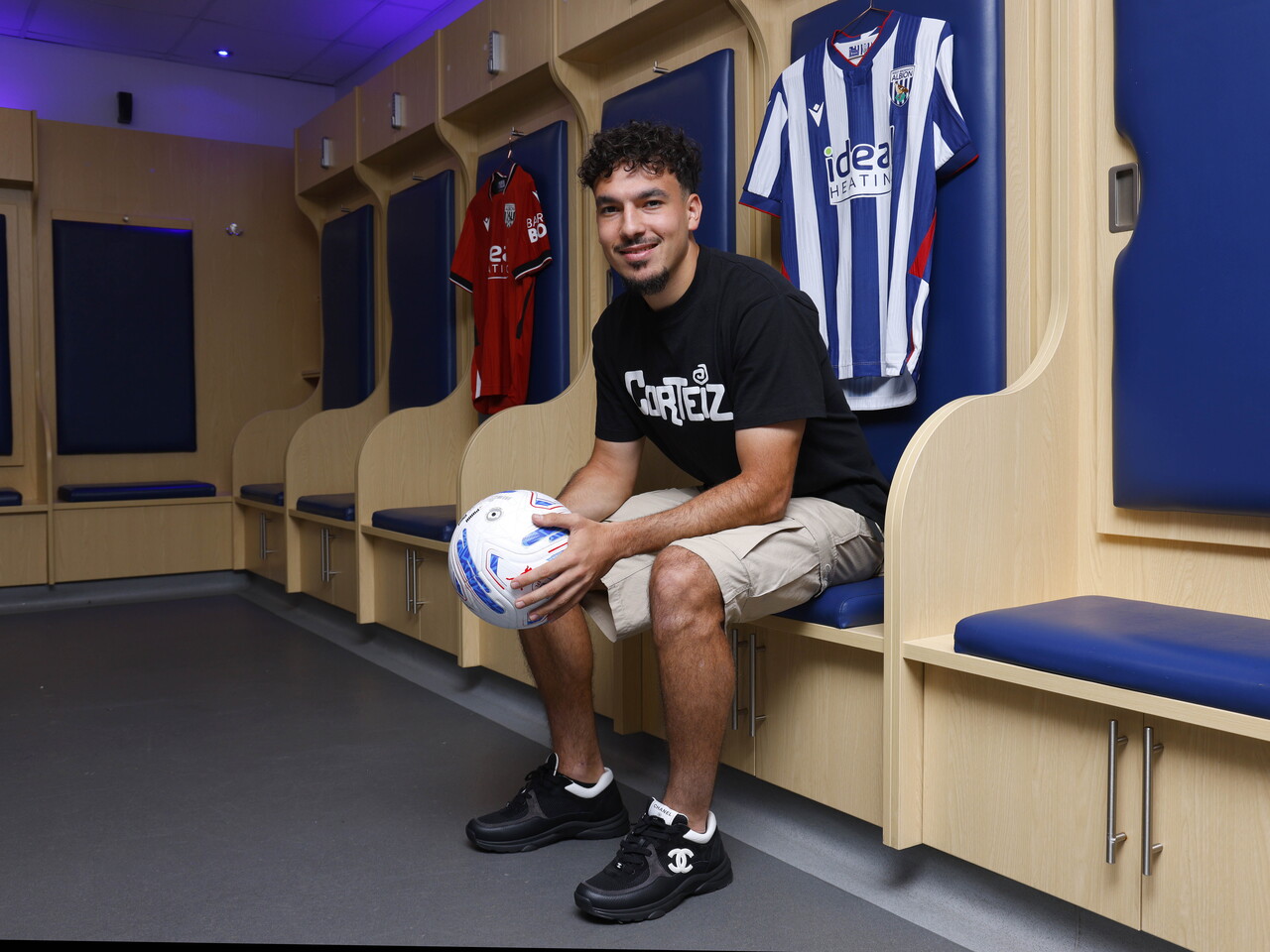 Nolan Galves sat in the home dressing room holding a ball smiling at the camera with a home shirt and an away shirt hanging up behind him