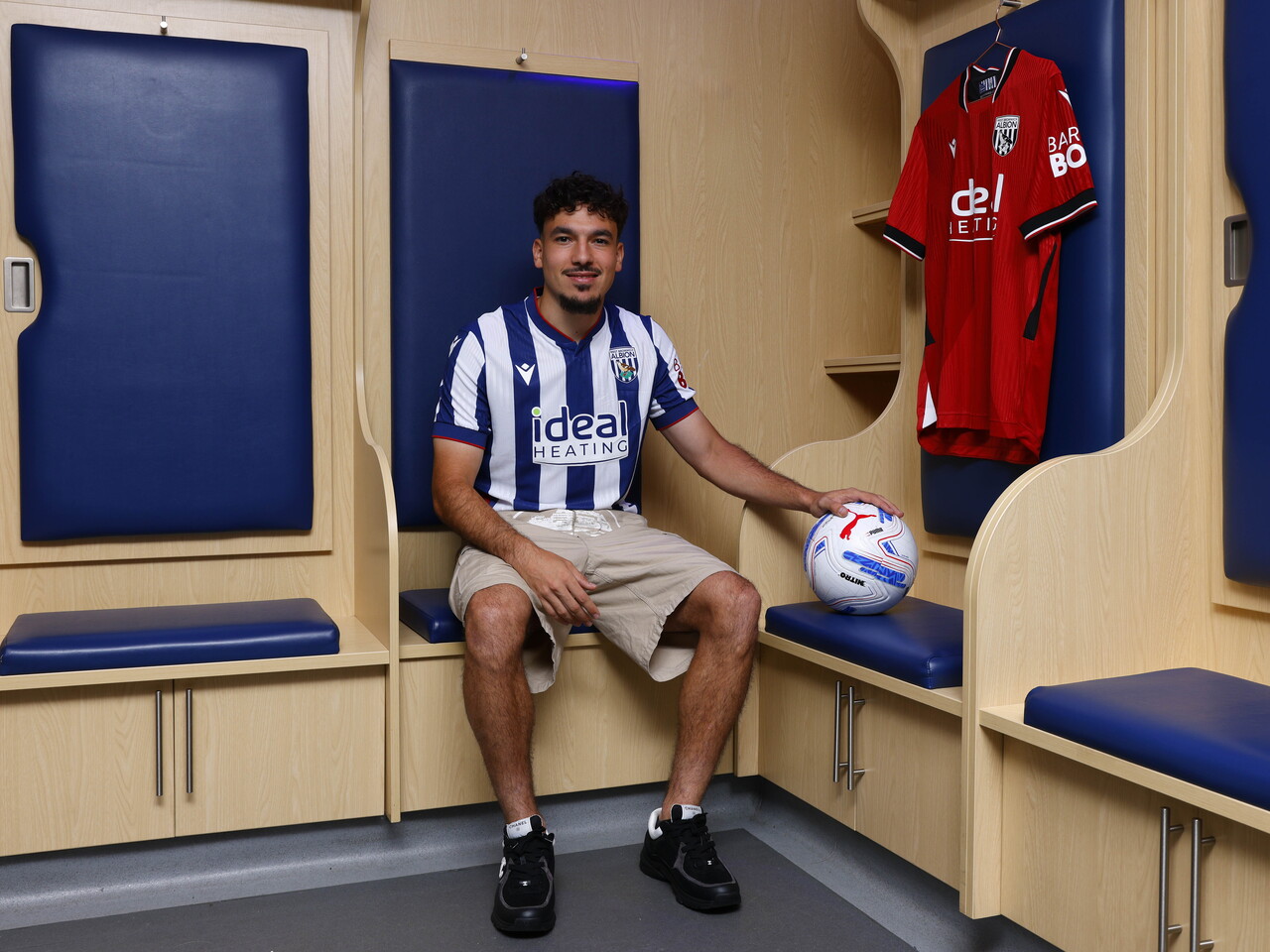 Nolan Galves sat in the home dressing room in the home kit holding a ball, with an away shirt hanging up behind him