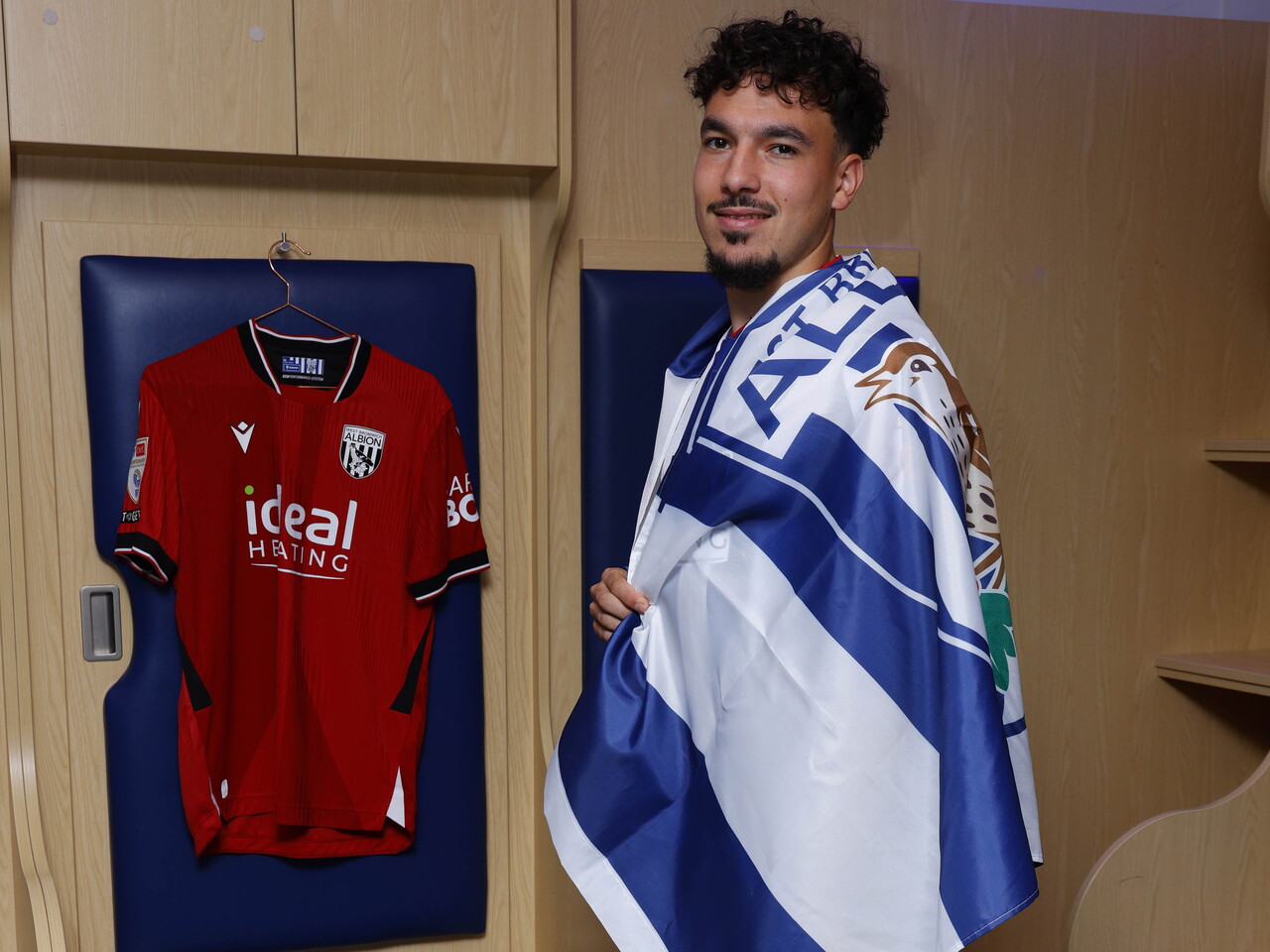 Nolan Galves in the home dressing room dressed in a WBA flag with an away shirt hanging up behind him