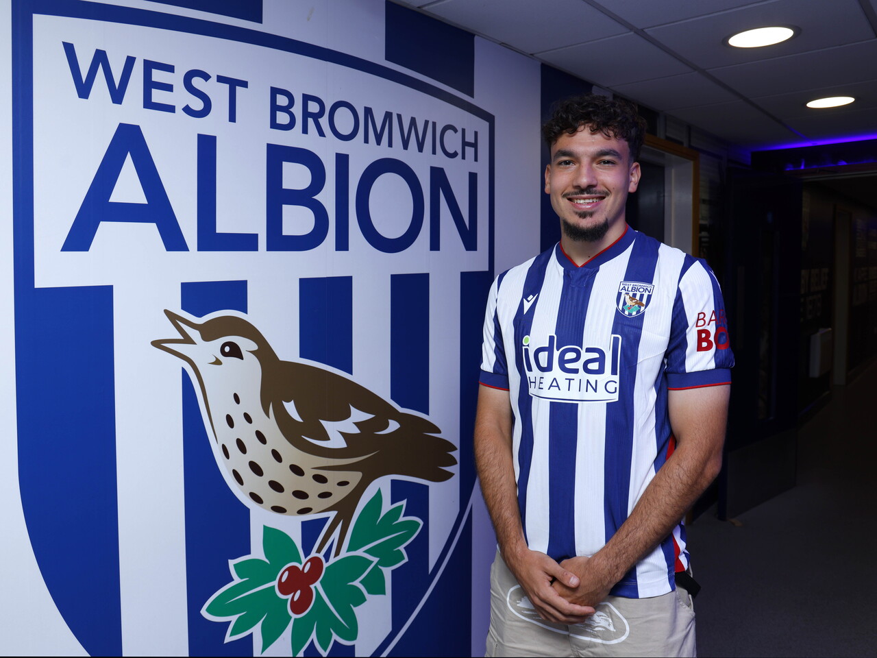 Nolan Galves smiling at the camera while stood next to a big WBA badge in the home shirt 