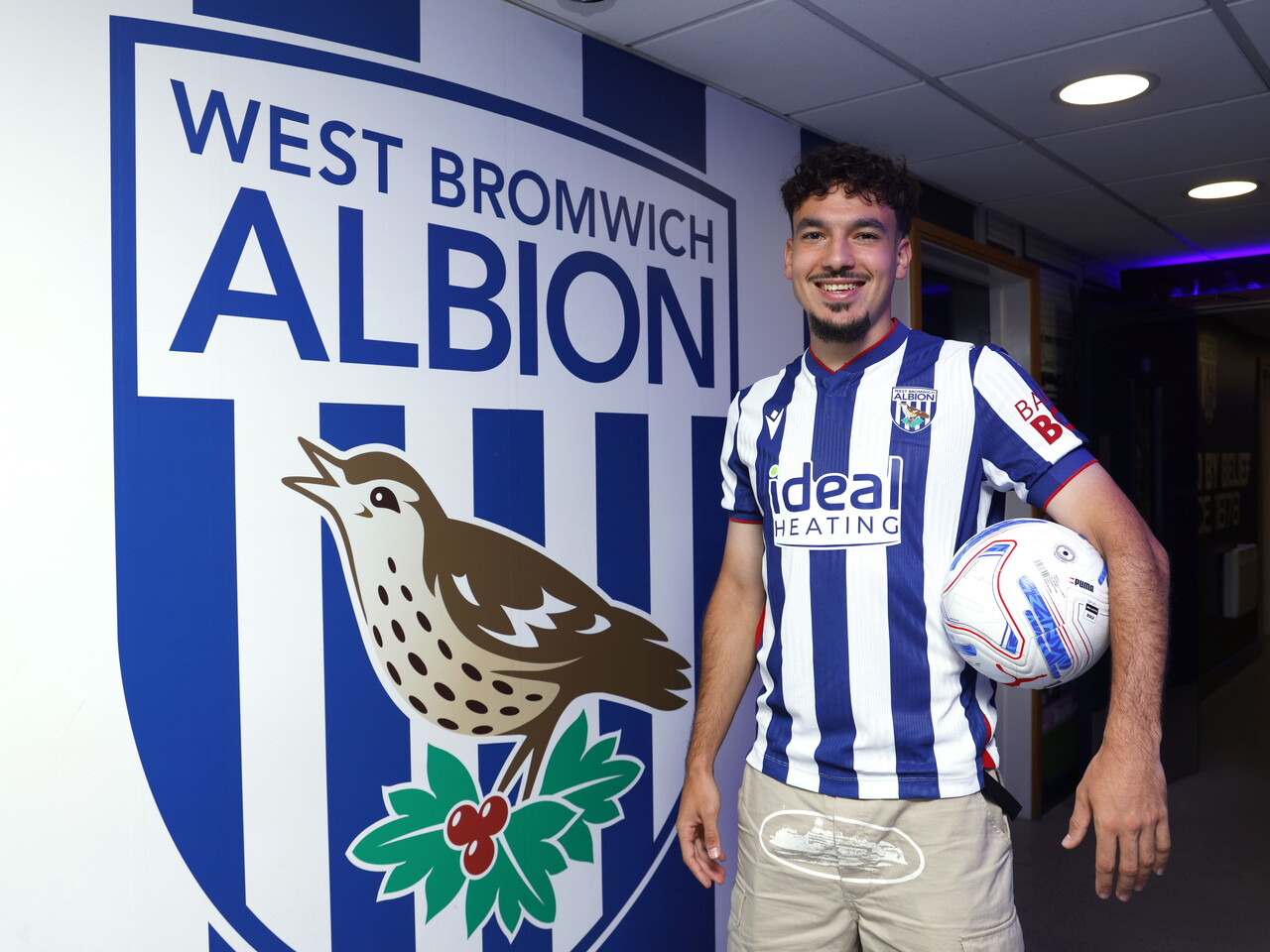 Nolan Galves smiling at the camera while stood next to a big WBA badge in the home shirt, while holding a ball