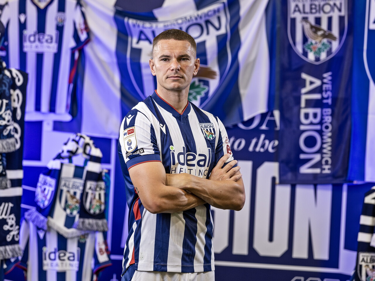 Conor Townsend in the home kit looking at the camera with his arms folded while stood in front of an Albion back drop with plenty of WBA branding