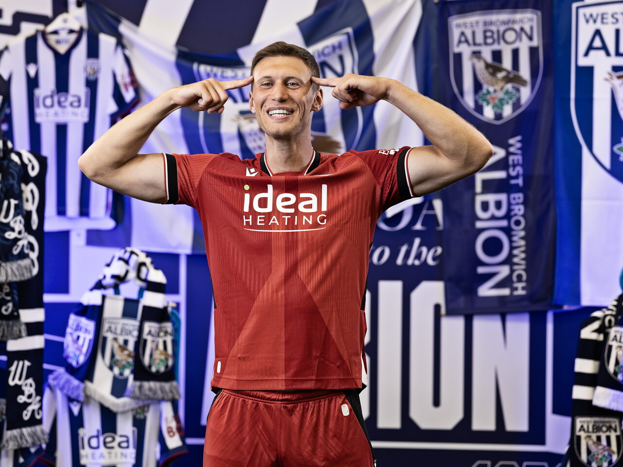 Krystian Bielik in the red away kit posing for the camera while stood in front of an Albion back drop with plenty of WBA branding