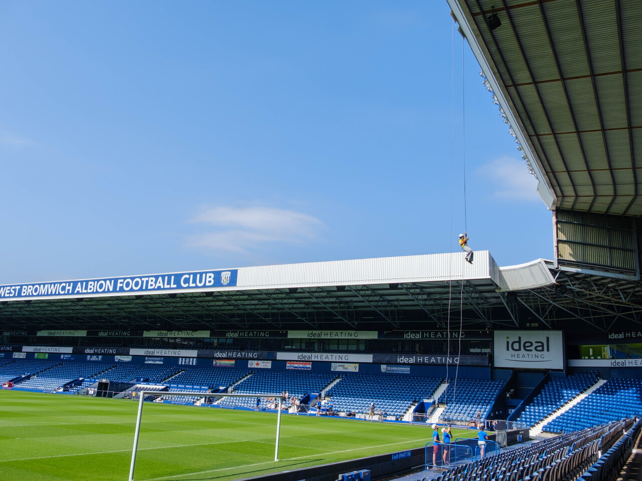 Participant abseils from the roof of The Hawthorns high above the West Bromwich Albion pitch, with blue seating and stadium stands visible below.