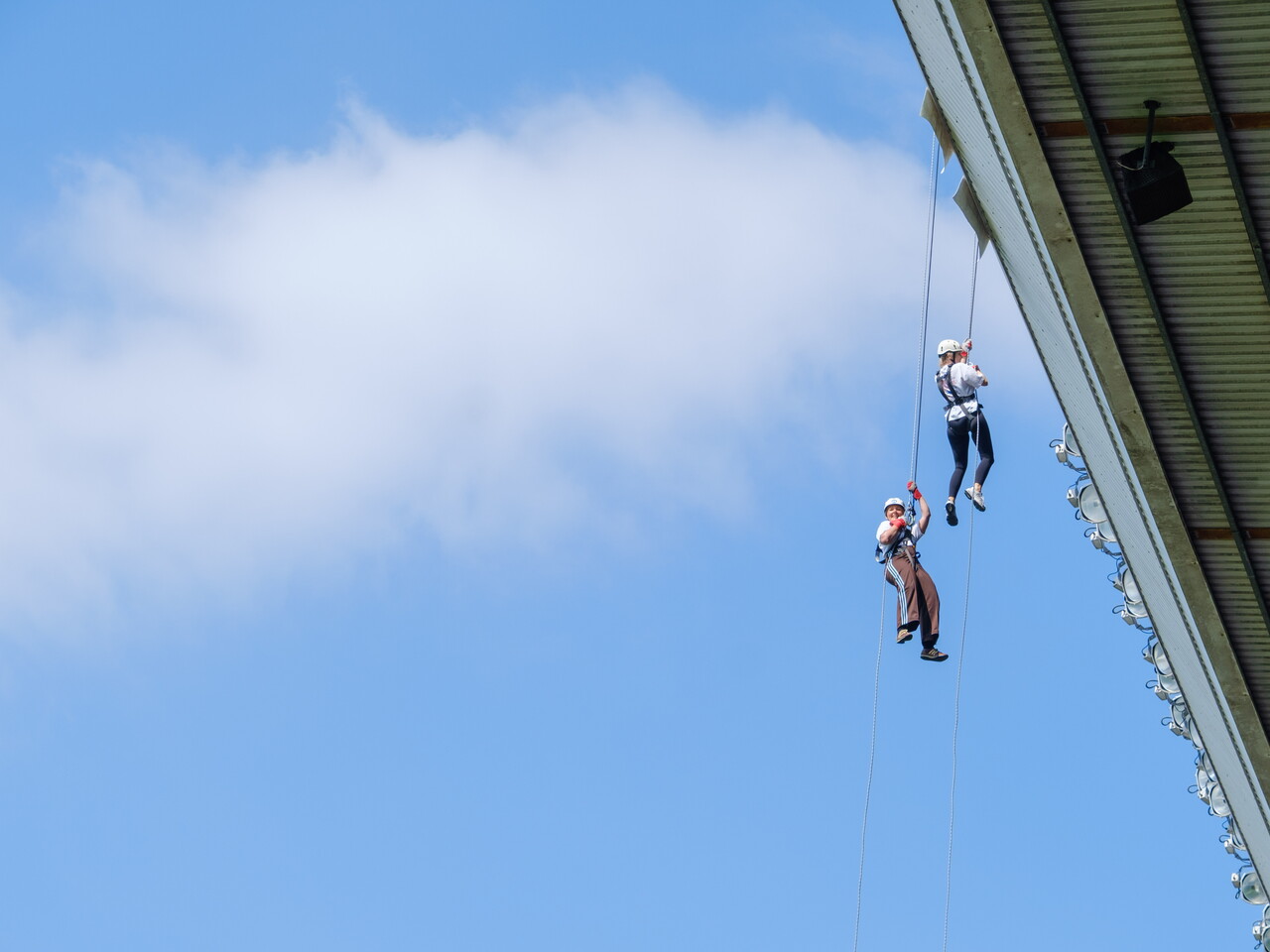 Two participants abseil side by side from the roof of The Hawthorns, suspended high above the stadium against a blue sky.