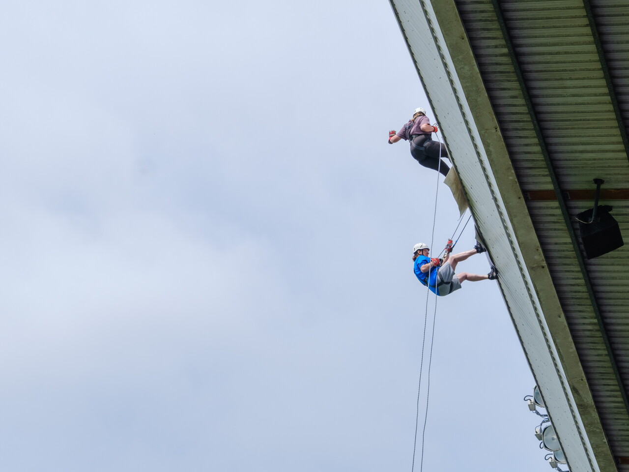 Two participants abseil side by side from the roof of The Hawthorns, suspended on ropes against the stadium structure and sky.