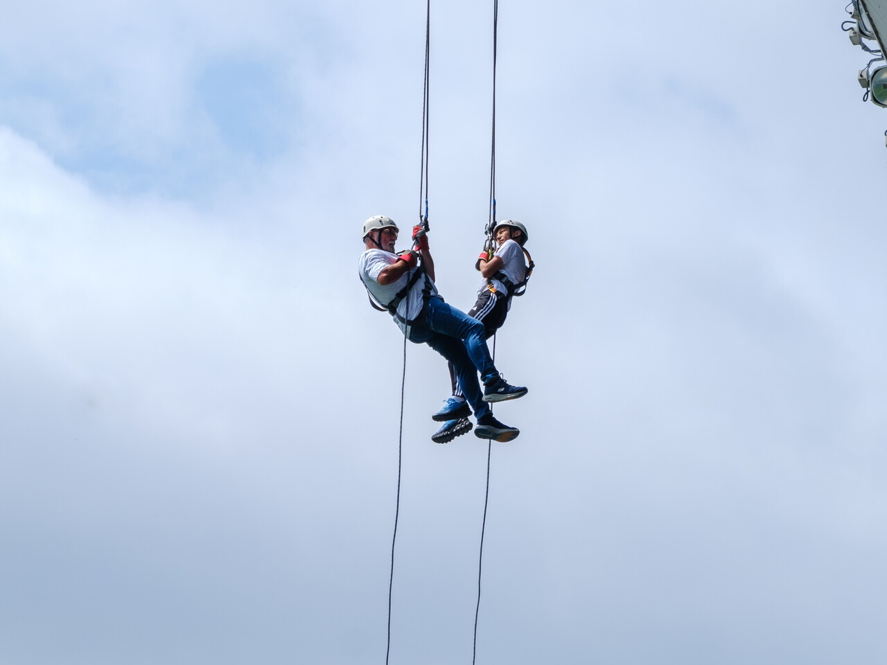 Two participants descend on ropes during a tandem abseil from the roof of The Hawthorns, suspended against a cloudy sky.