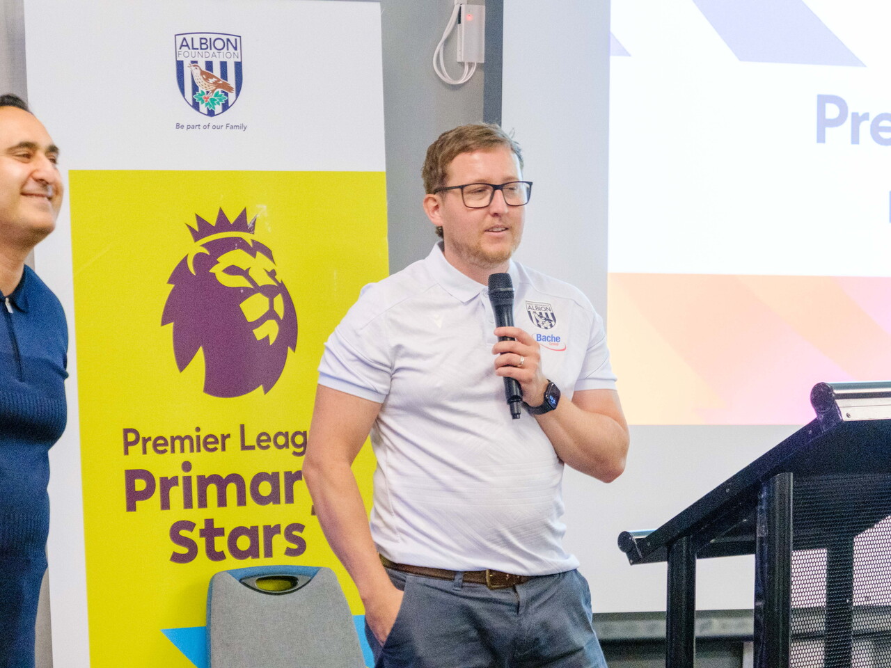 Deputy Director Jon Ward speaking on the mic at the front of the room with Rupi Lal at the side. 