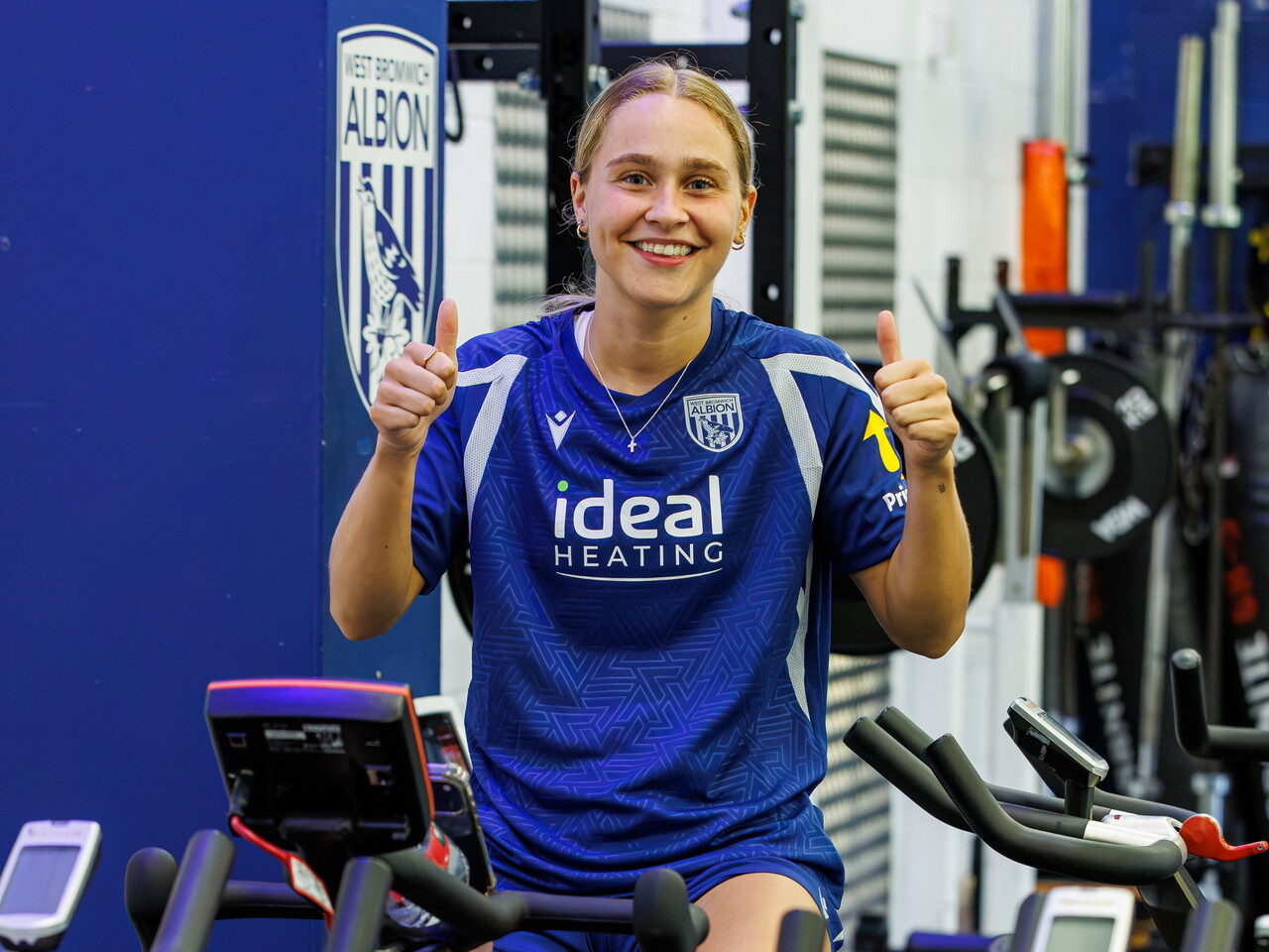 Mia Sorrentino sat on a bike in the gym smiling at the camera with her thumbs up
