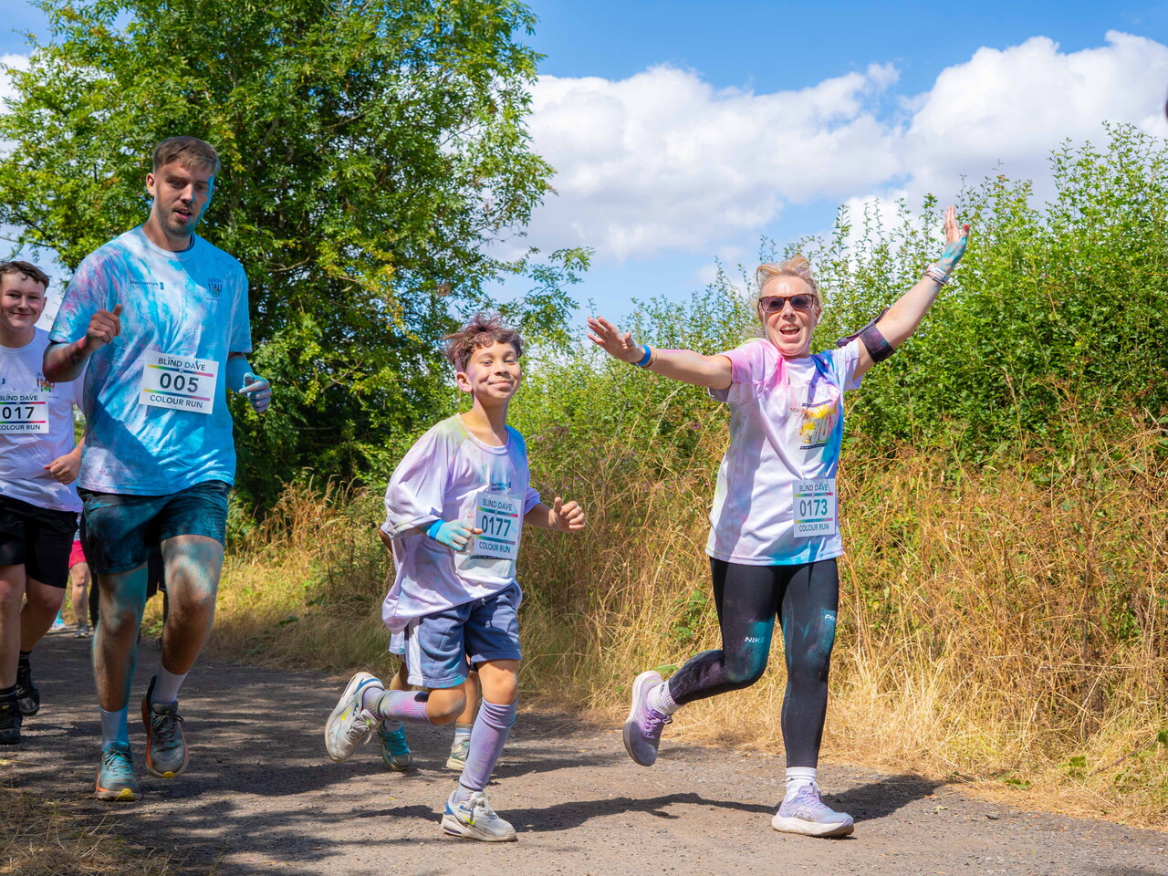 Participants running and smiling along a path tall grass and trees.