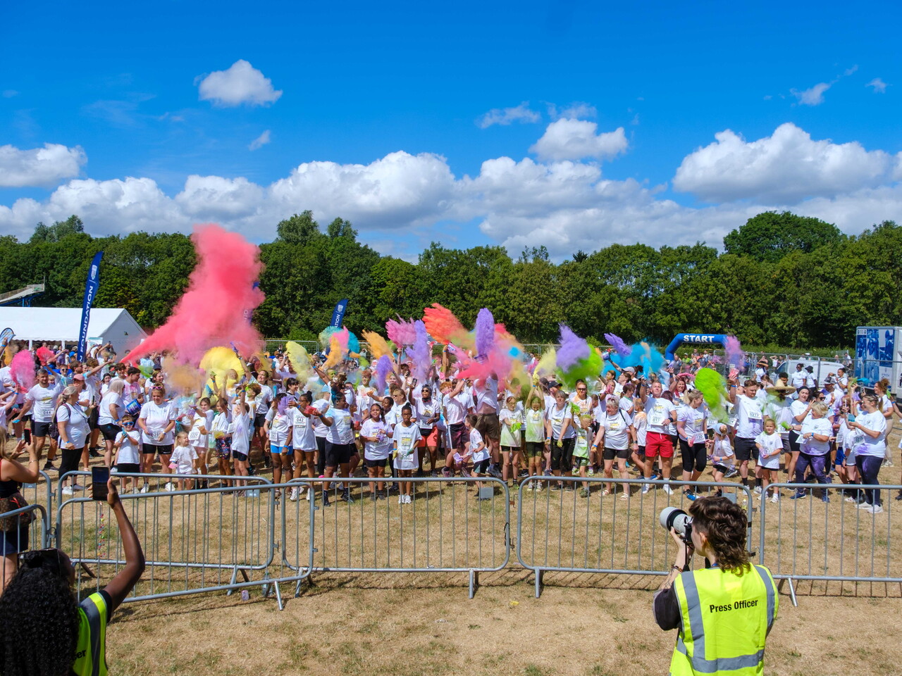 Participants throw brightly coloured powder into the air during a charity colour run, creating large clouds of pink, purple, blue, yellow and green across an open field.