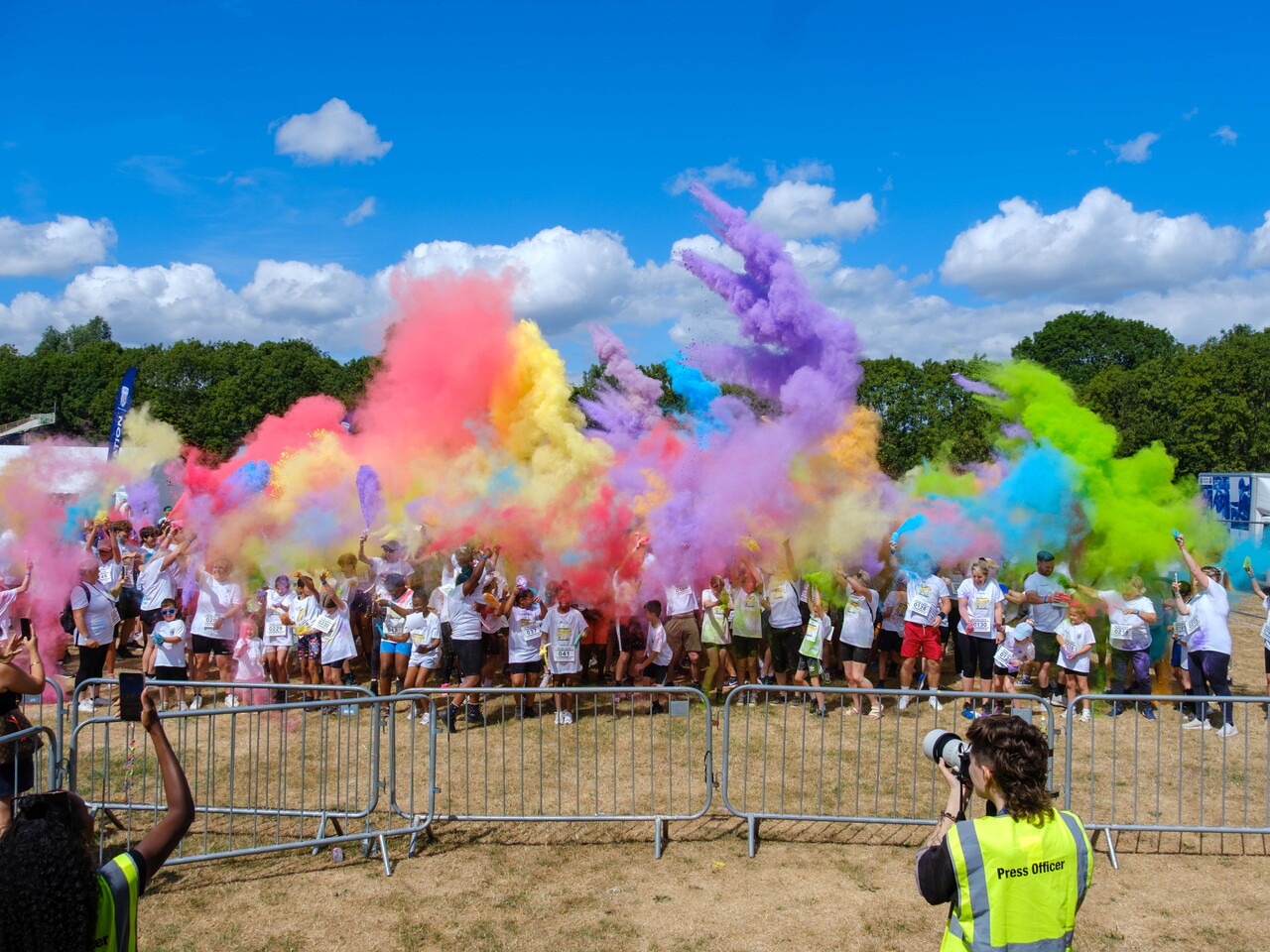 Participants throw brightly coloured powder into the air during a charity colour run, creating large clouds of pink, purple, blue, yellow and green across an open field.
