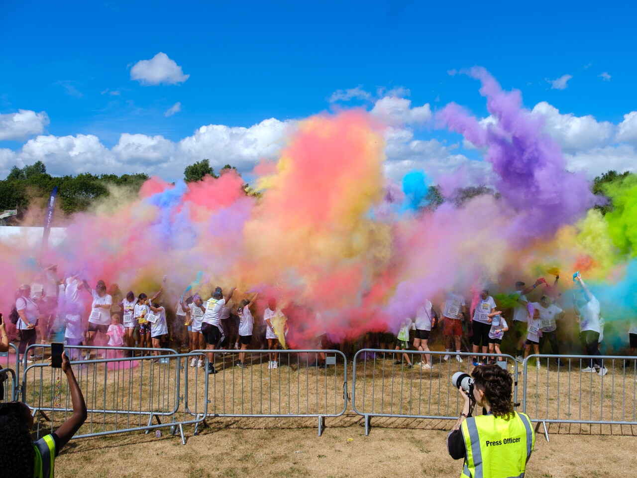 Participants throw brightly coloured powder into the air during a charity colour run, creating large clouds of pink, purple, blue, yellow and green across an open field.