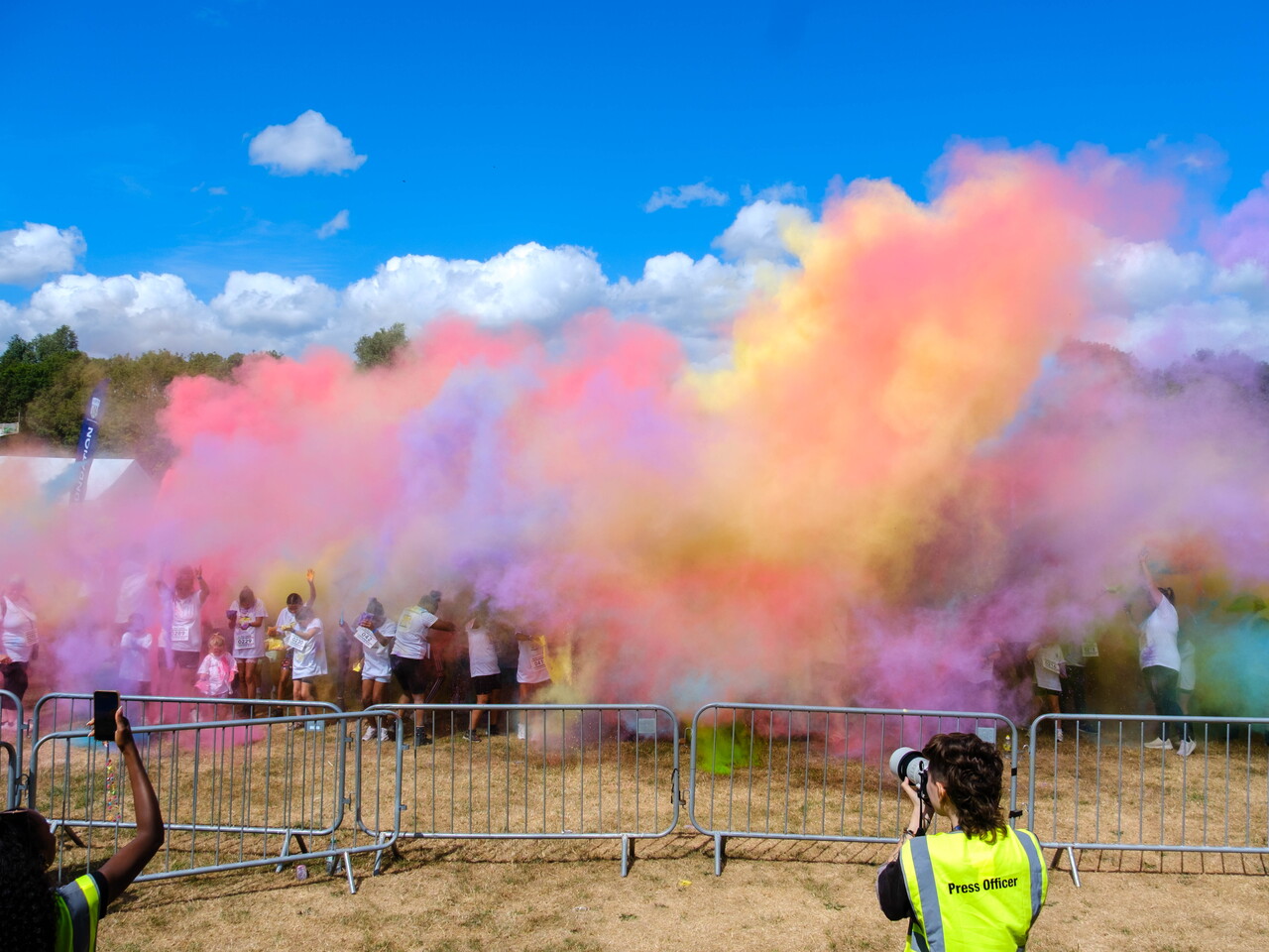 Participants throw brightly coloured powder into the air during a charity colour run, creating large clouds of pink, purple, blue, yellow and green across an open field.