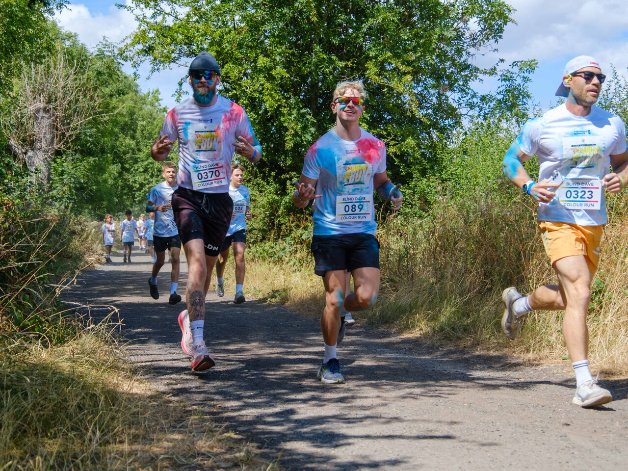 Several participants covered in blue and pink color powder run along a tree-lined countryside path