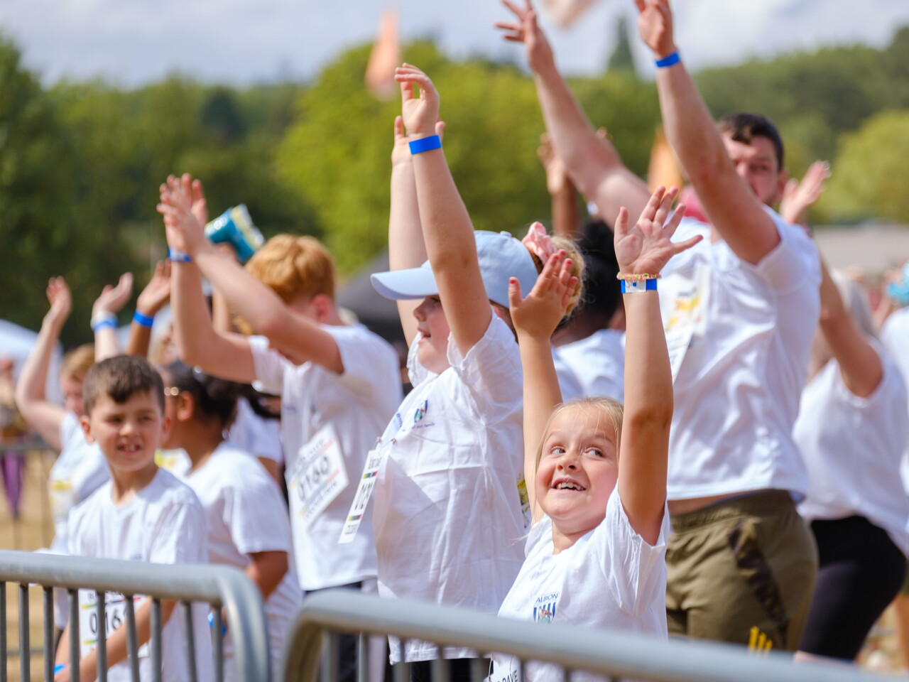 Colour run participants raise their hands during a warm-up session behind barriers, wearing event shirts and wristbands before the start of the run.