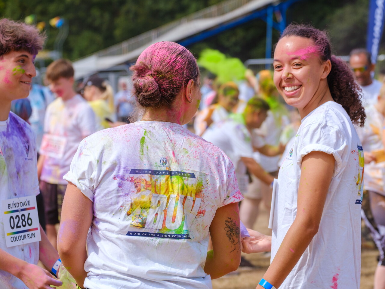 Colour run participants covered in brightly coloured powder gather on an open field, wearing event shirts and bibs during the Blind Dave Colour Run.