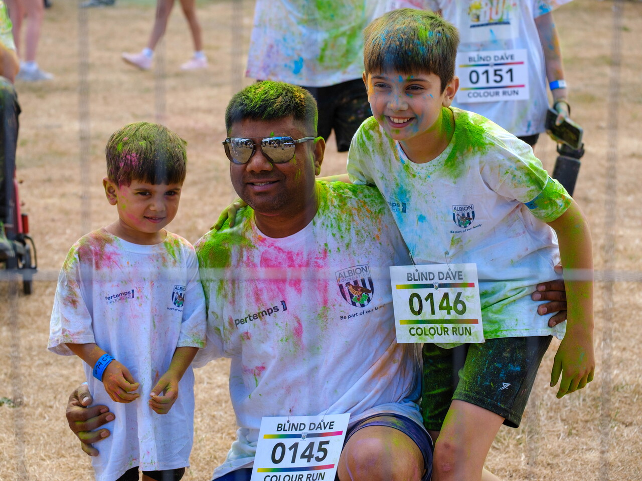 Adult and two children pose together after the Blind Dave Colour Run, covered in coloured powder and wearing event shirts and numbered bibs.