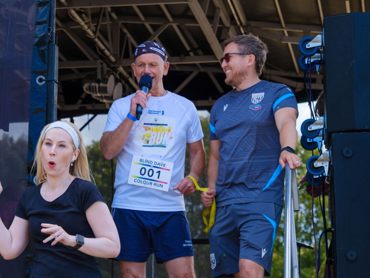 Blind Dave speaks on stage with a microphone before the charity colour run, alongside an Albion Foundation representative, while a British Sign Language interpreter stands in front of the stage.