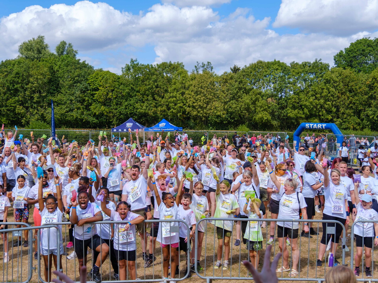 Large group of charity colour run participants gather behind barriers before the start, raising packets of coloured powder in the air on a sunny field, with event tents and a START arch visible in the background.