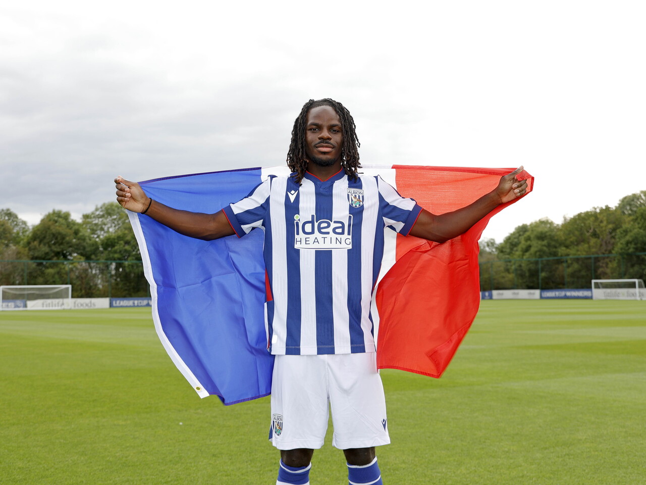 Brayann Pereira smiling at the camera while holding a French flag and stood in the home kit on a training pitch