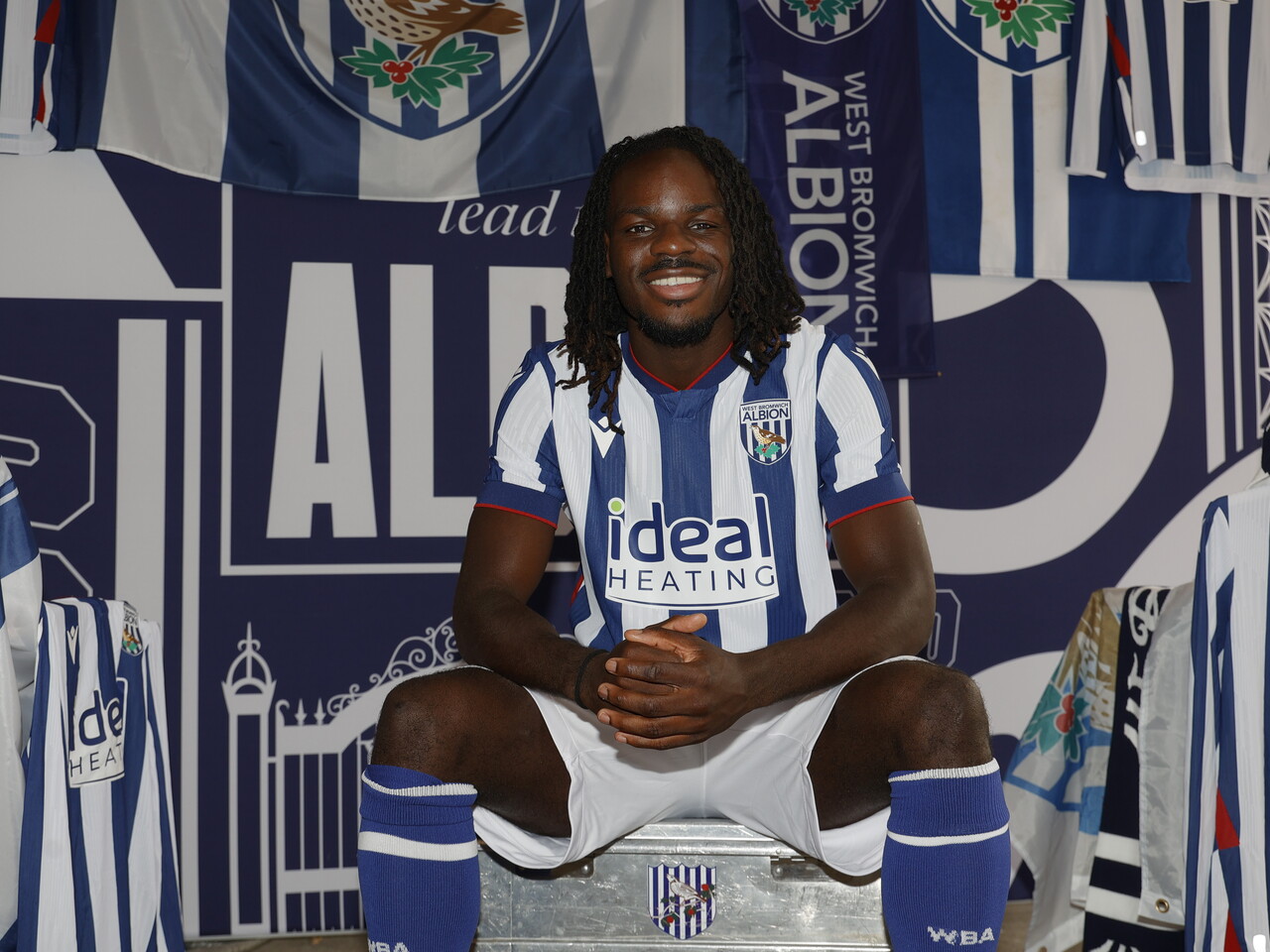 Brayann Pereira smiling at the camera while sat on a box in the home kit in front of WBA branding 