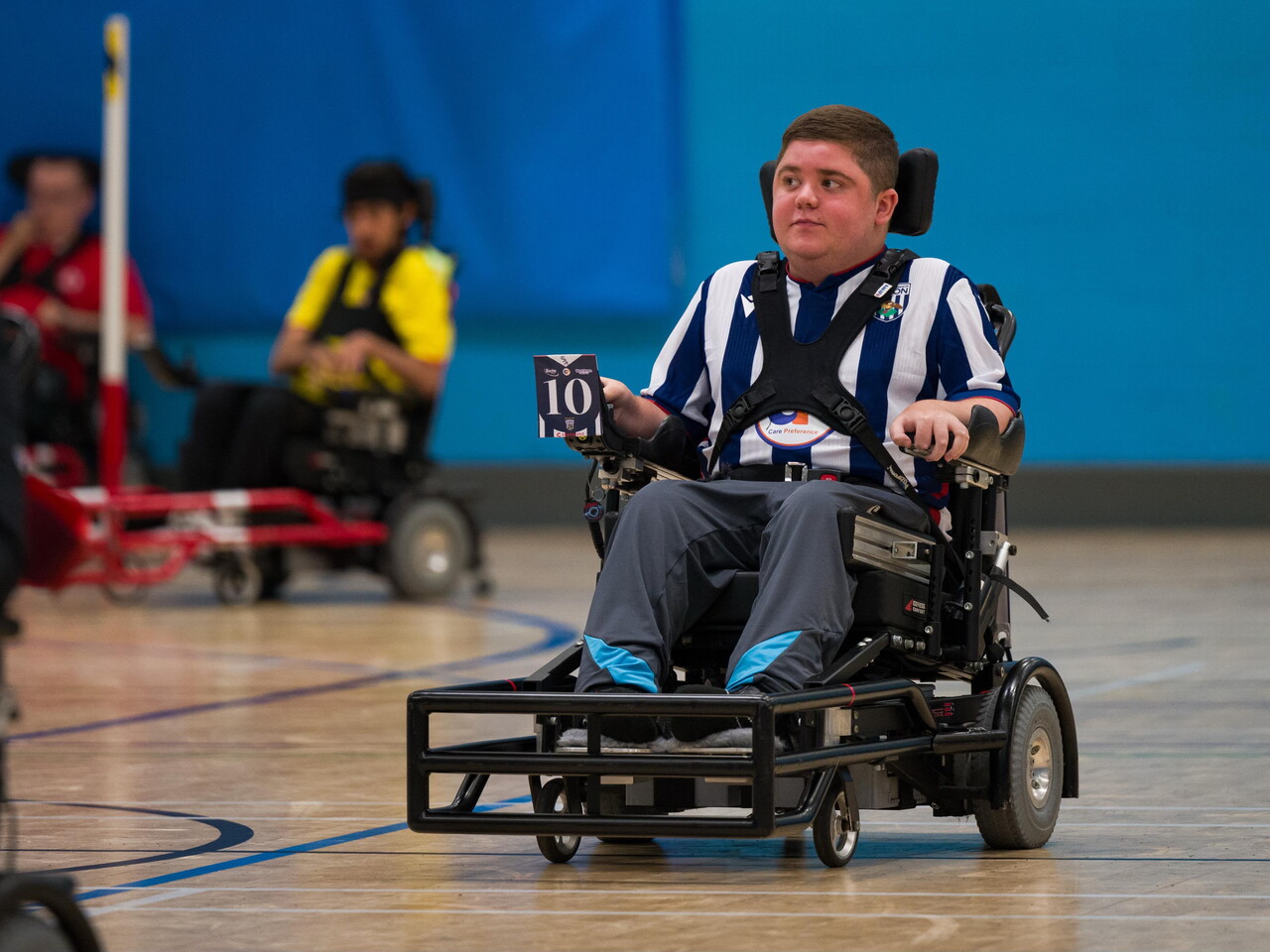Buch-Platt, wearing a blue-and-white striped West Bromwich Albion Powerchair Football kit, drives a powerchair across an indoor court during a match, with other players visible in the background.