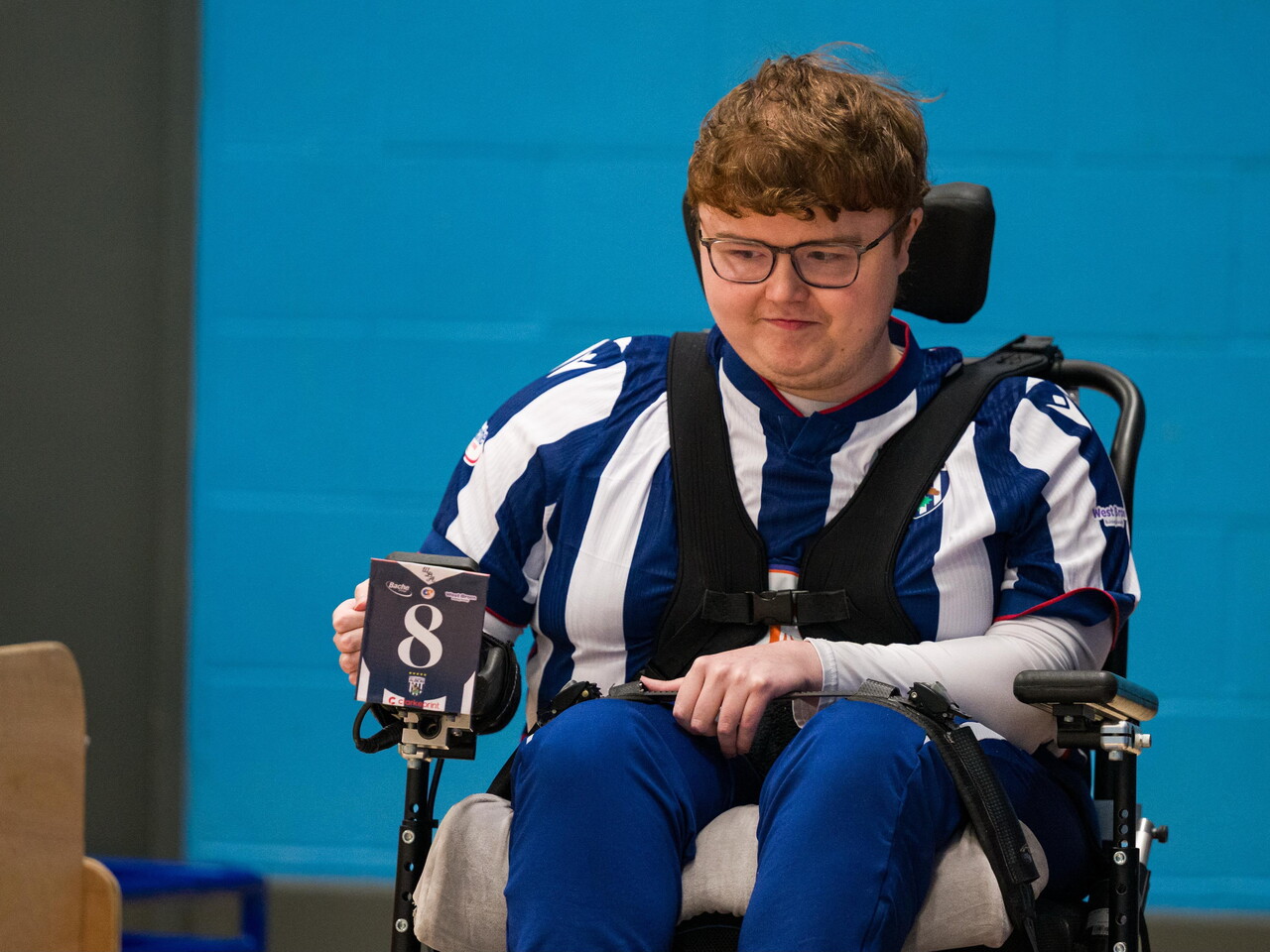 Christer, wearing a blue-and-white striped West Bromwich Albion Powerchair Football kit, sits in a powerchair during an indoor match, with a player number card attached to the chair.