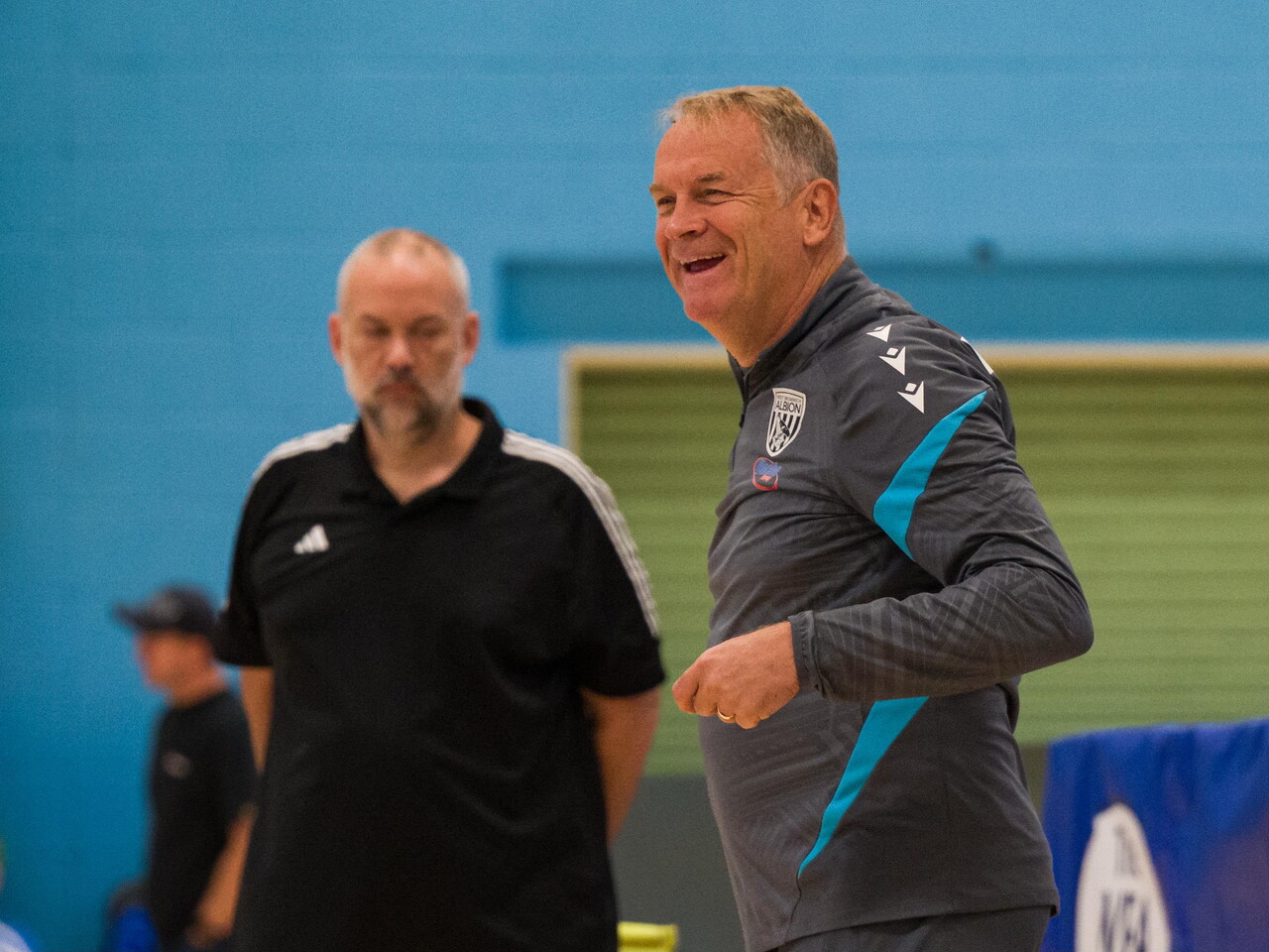 Coach Colin Gordon stands courtside during an indoor powerchair football match, observing play in a sports hall.