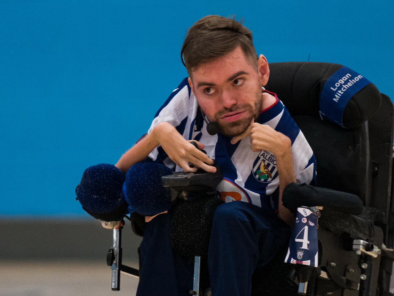 Harrison, wearing a blue-and-white striped West Bromwich Albion Powerchair Football kit, controls a powerchair during an indoor match, with a player number card attached to the chair.