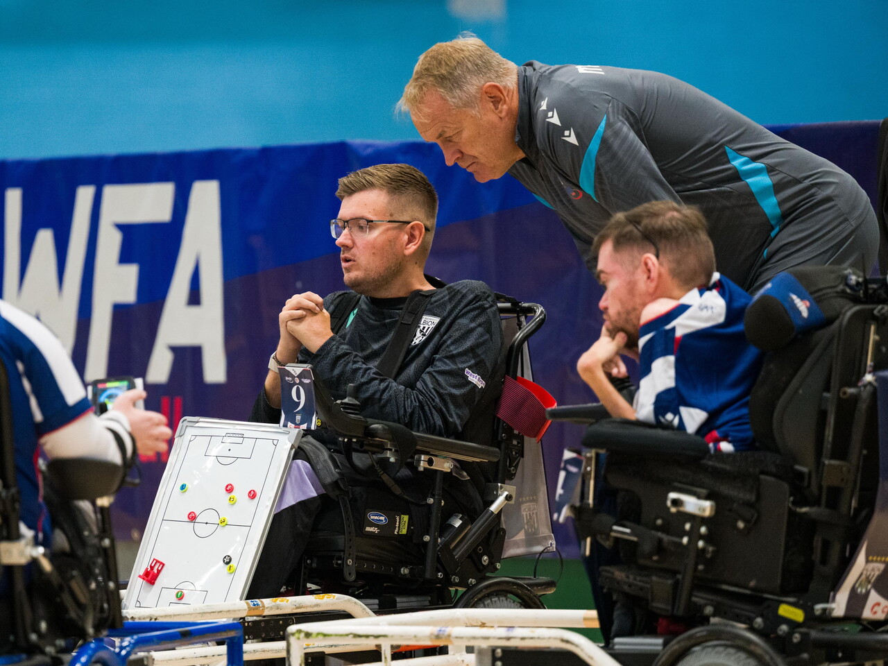 Player-coach Chris Gordon discusses tactics with teammates from a powerchair, while Coach Colin Gordon leans over to contribute during a break in play.