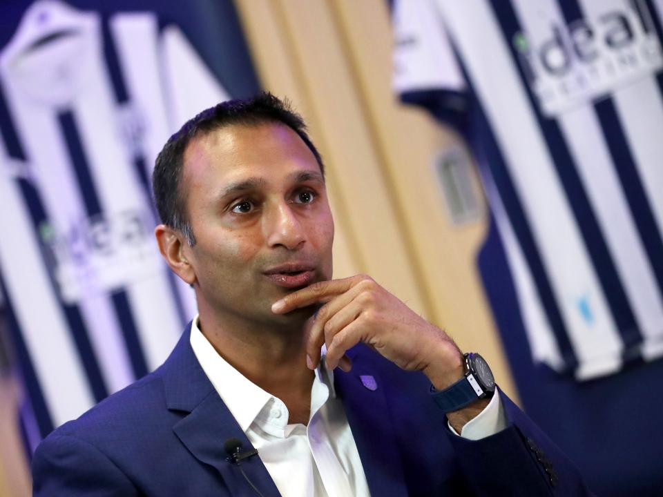 Shilen Patel facing forward with his finger on his chin with two Albion shirts hanging up behind him