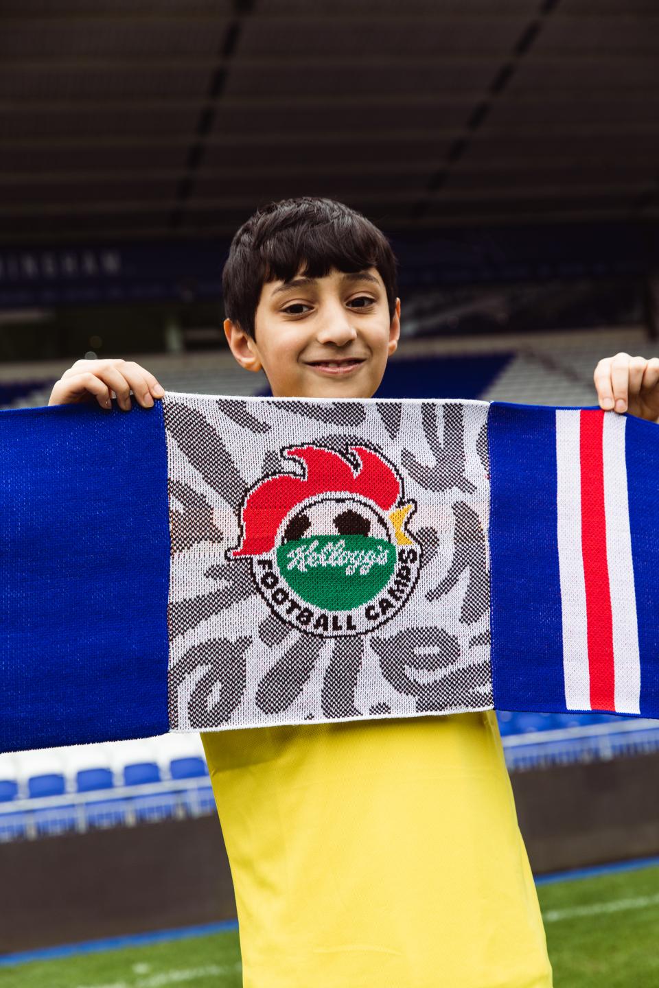 Child holding a blue, white & red scarf with Kelloggs Football Camps branding in the centre.