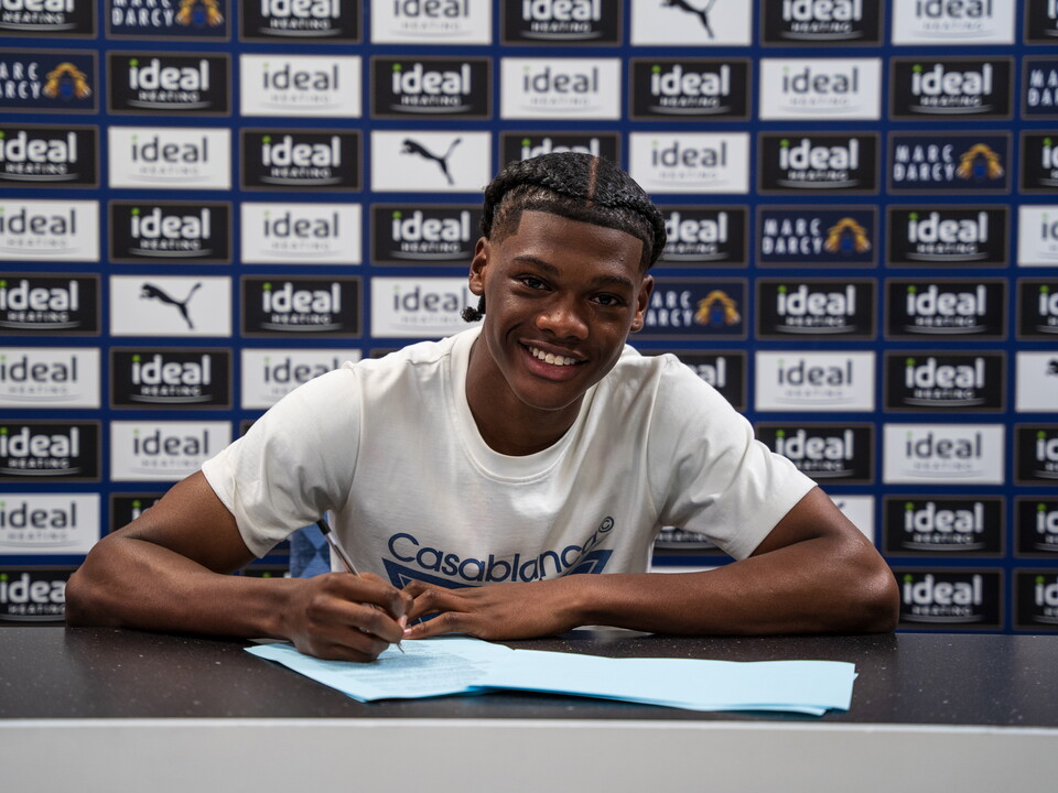 A photo of academy graduate Adriel Walker signing his professional contract in front of a sponsors billboard at the training ground