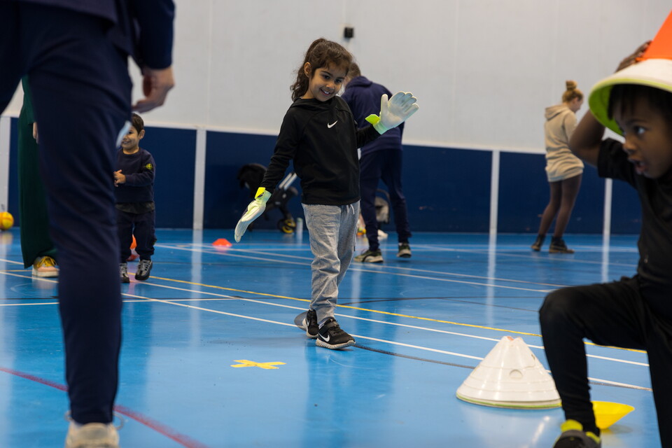 Khadija walks forwards on the blue sports hall surface wearing her goalkeeper gloves.