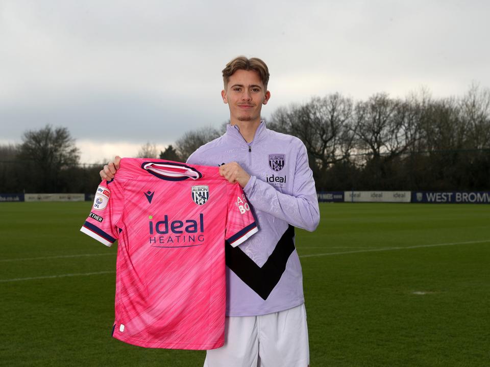 Isaac Price holding up a pink WBA shirt and smiling at the camera 