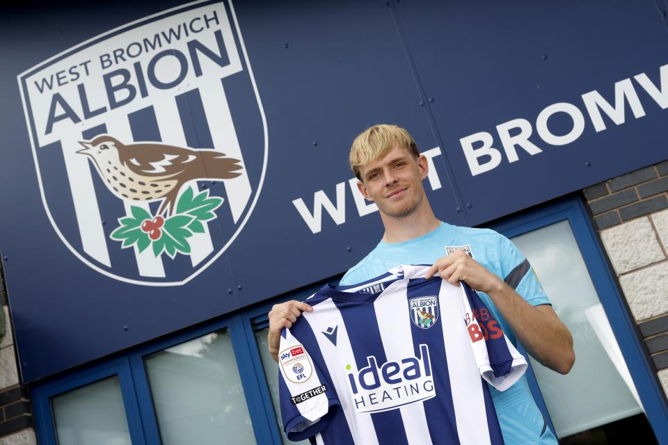 Toby Collyer holding up a WBA home shirt and smiling at the camera 
