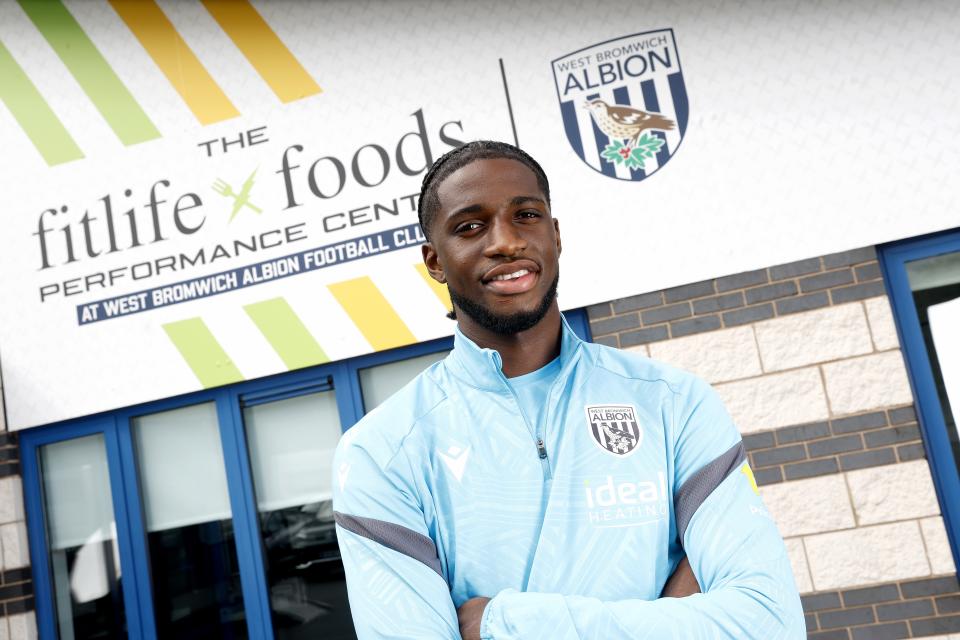 Samuel Iling-Junior smiling at the camera while stood in front of branding outside the training ground 