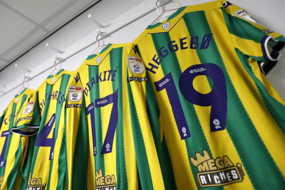 Several green and yellow WBA away shirts hanging up in the dressing room with names and numbers facing the camera 