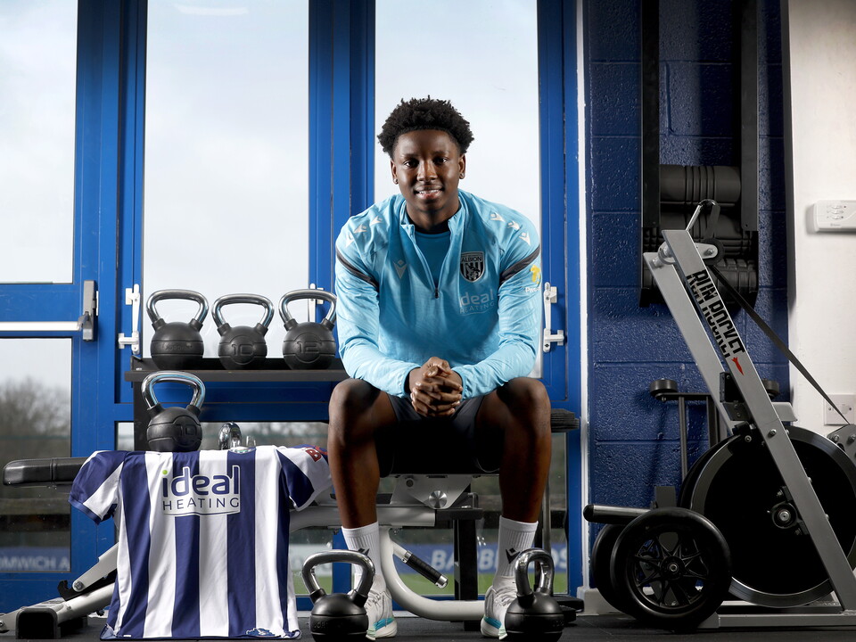 Jamaldeen Jimoh-Aloba smiling at the camera while sat in the gym on a bench with a home shirt next to him