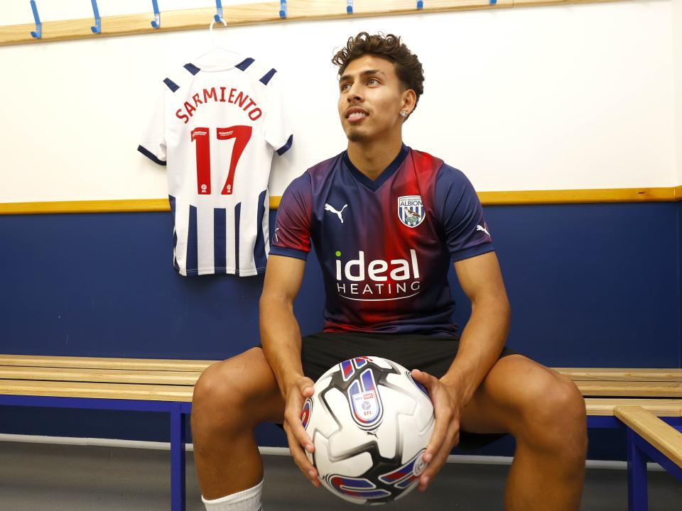 Jeremy Sarmiento looks away from camera while holding a ball with his shirt hanging up in the background